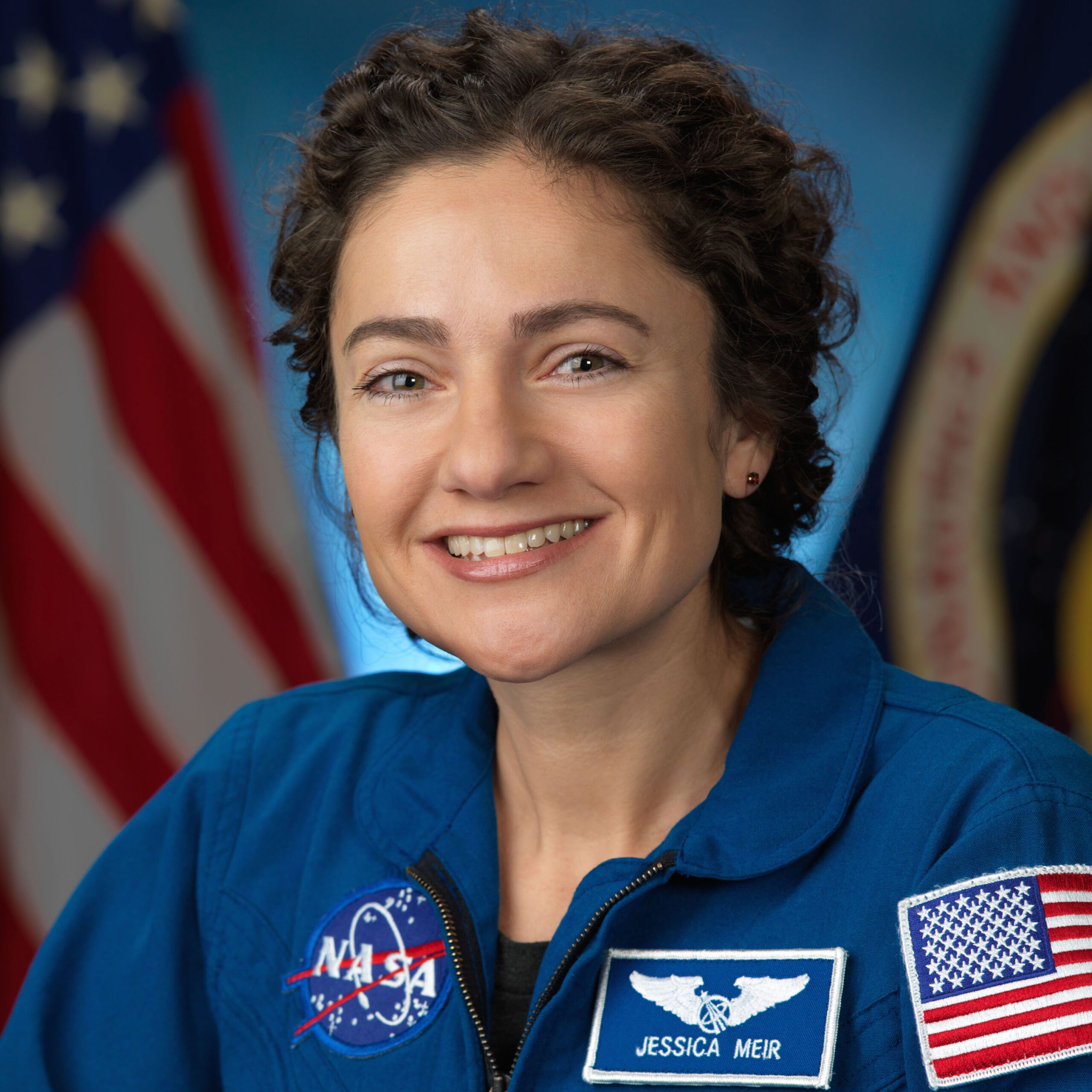 A smiling woman in a blue astronaut suit with badges.