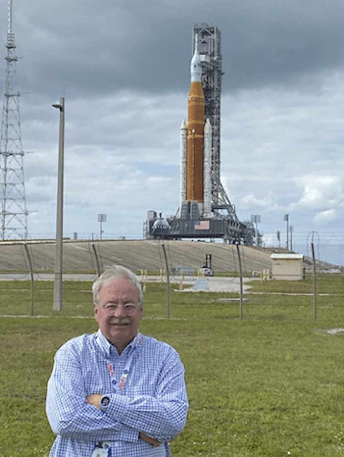Man smiling in front of a large rocket on a launch pad.