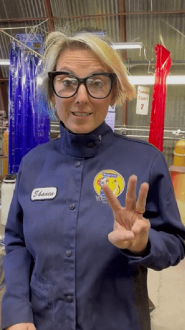 Person in safety gear holding up three fingers in welding workshop.