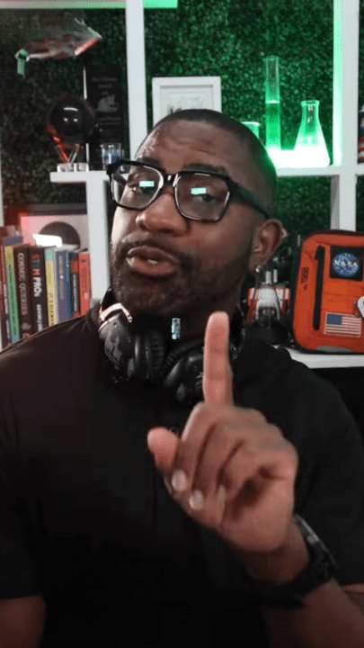 Person wearing glasses and headphones, gesturing, with books and lab equipment in the background.