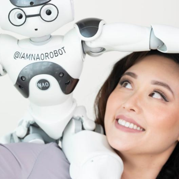 A woman smiling at a small, humanoid robot resting on her shoulder.