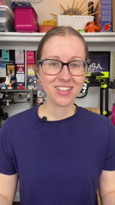 Person in glasses smiling, standing in front of a tech-filled workspace