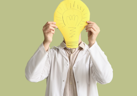 Person in lab coat holding a large paper light bulb over face.