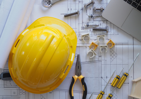 Engineering tools and yellow hard hat on blueprint, laptop nearby.
