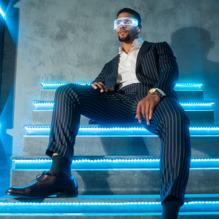 Man in pinstripe suit sitting on LED-lit stairs wearing futuristic glasses.