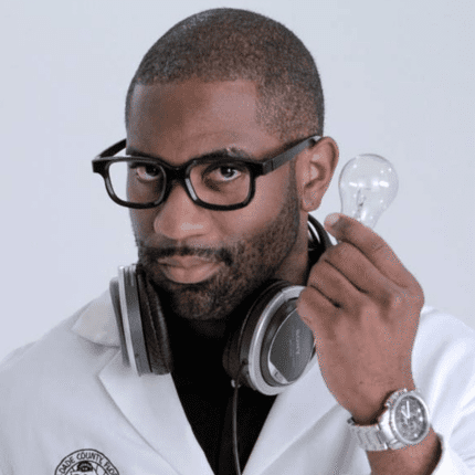 Man in glasses holding a light bulb, wearing a lab coat and headphones.
