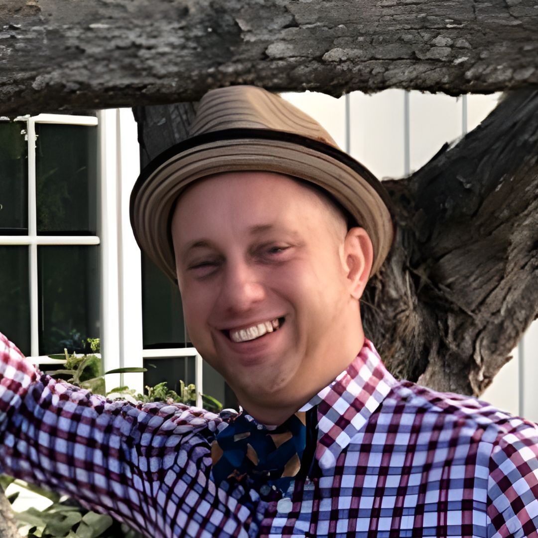 Smiling man wearing a checkered shirt and fedora hat.