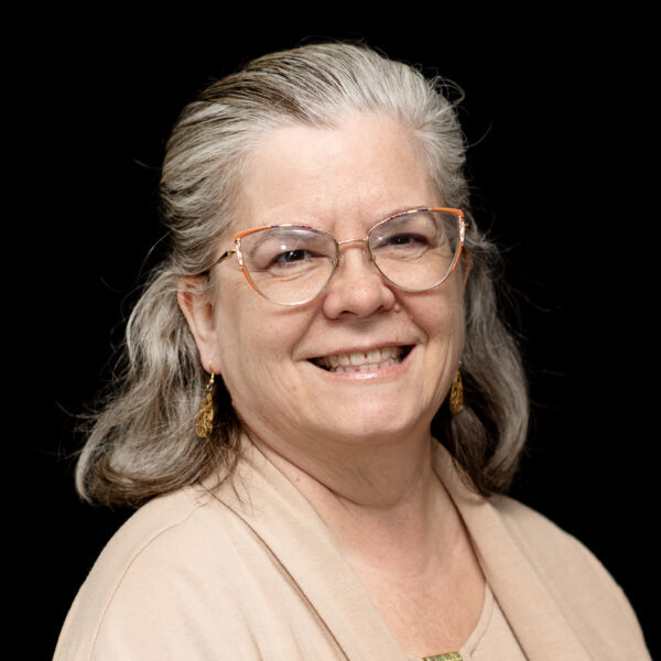 Portrait of a smiling woman with glasses and gray hair against a black background.